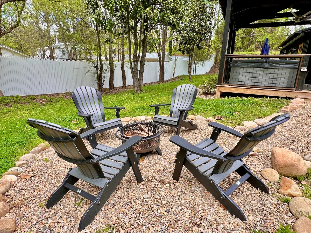 A cozy fire pit area featuring four black Adirondack chairs arranged on a pebbled ground surrounded by lush grass and trees. The setup invites guests to gather for evening stories, roast marshmallows, and enjoy the warmth of the fire under the stars.