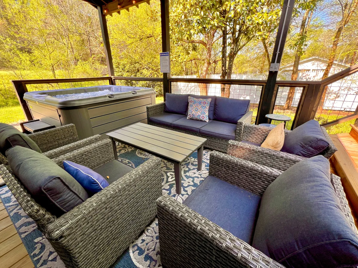 A serene outdoor deck with a bubbling hot tub, plush seating adorned with decorative pillows, and a coffee table, all surrounded by lush greenery. The area exudes comfort and invites guests to relax, socialize, or enjoy a morning coffee while soaking up the view.