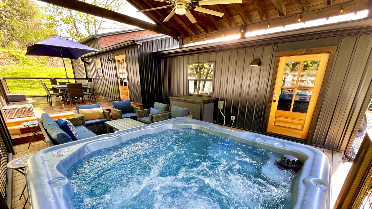 The image showcases a stylish indoor-outdoor space featuring a bubbling hot tub, inviting seating area, rustic wooden accents, and an outdoor dining set—all with views of the lush Smoky Mountains just beyond. The space exudes warmth and relaxation, suggesting the perfect spot for unwinding or gathering with friends and family.