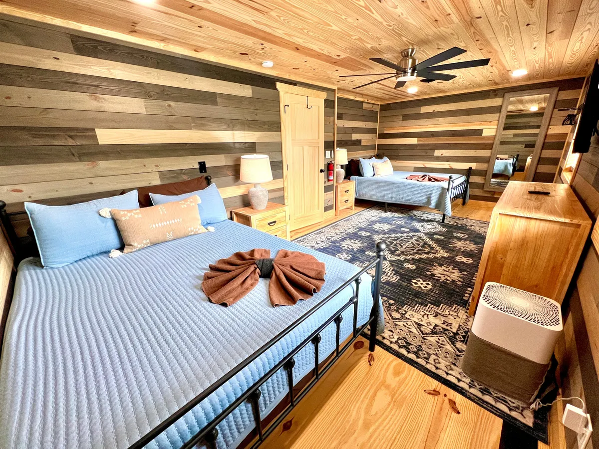 Two beds with light blue bedding rest against striped wooden walls. A large mirror and an area rug complete the space, providing a sense of openness and a welcoming environment ideal for families or groups.