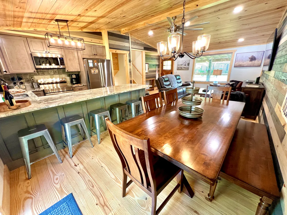 A wide-angle view of the dining table and kitchen island, with a chandelier overhead and comfortable living area seating nearby. The scene is bright and inviting, clearly showing the flow between kitchen, dining, and relaxing spaces.