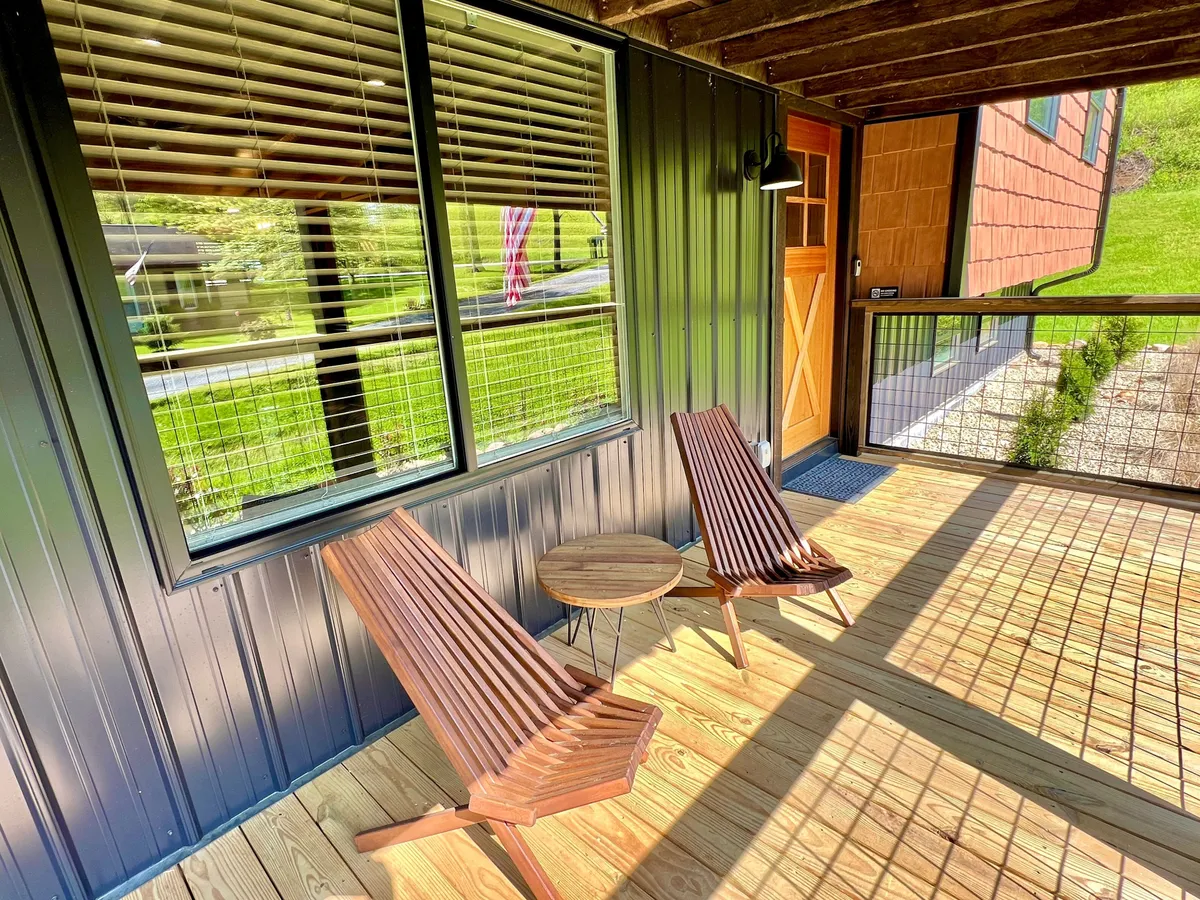 A serene outdoor scene featuring two wooden lounge chairs and a small round table on the deck, with a window and lush greenery in the background. The inviting setup suggests quiet mornings with coffee or intimate conversations, aligning with the idea of seclusion and relaxation in nature.