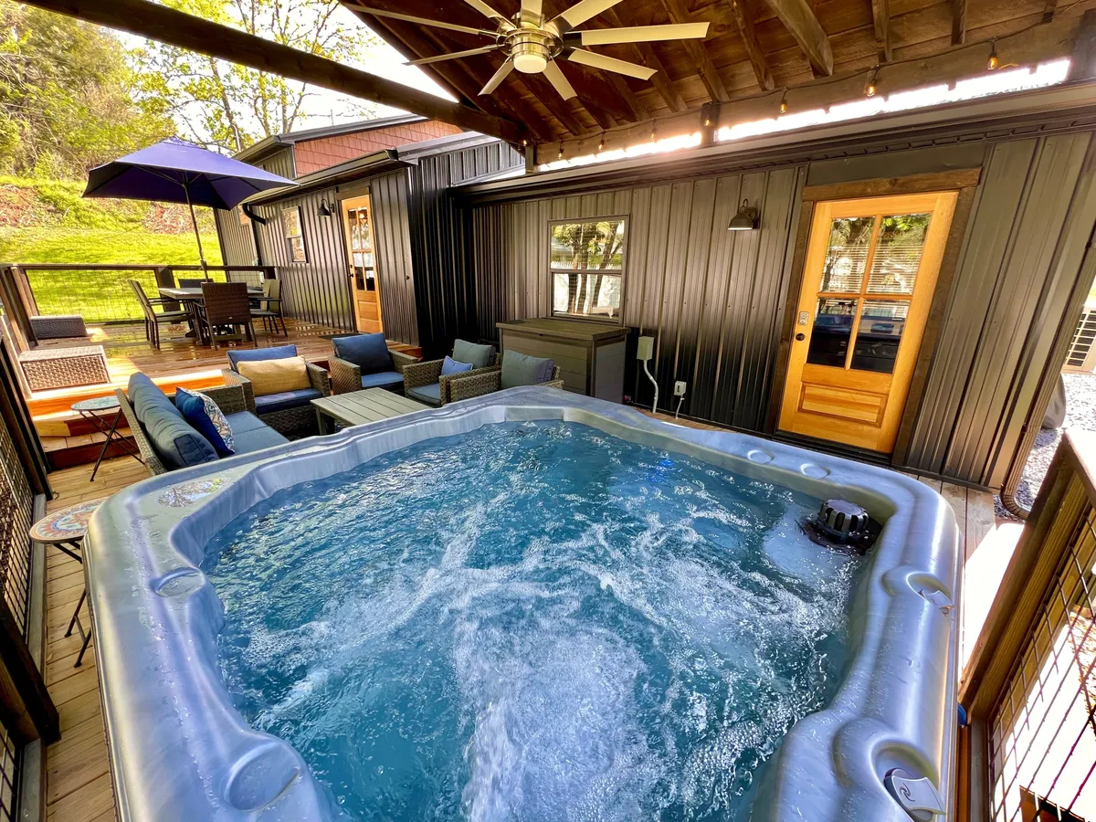 An inviting outdoor seating and hot tub area beneath a wooden ceiling with an umbrella, blending luxury with a cozy, resort-style atmosphere. The setup is intimate and designed for relaxation or entertaining, with the potential for starlit evenings and intimate dinners.