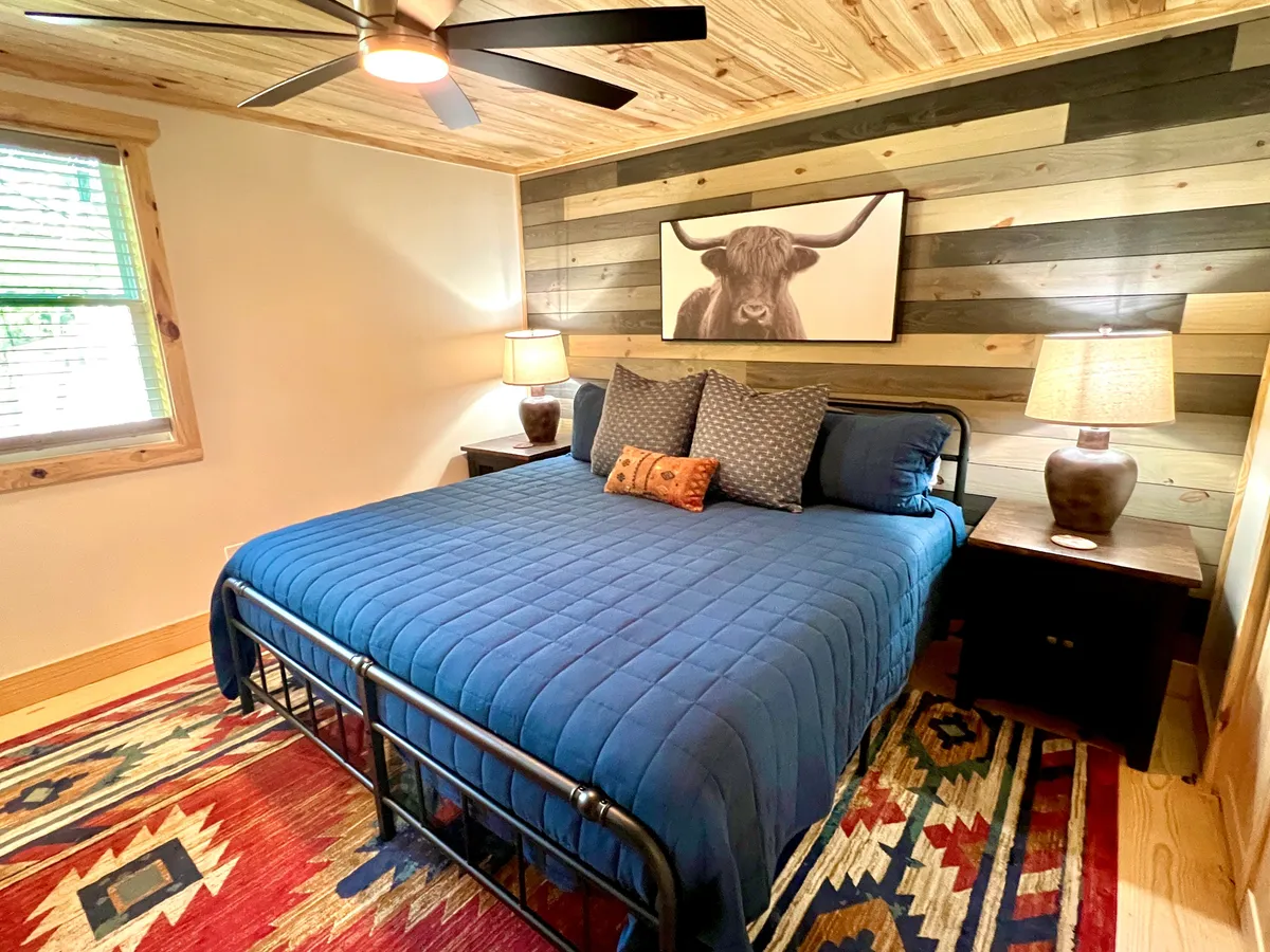 This image features a bed with a blue quilt, a wooden accent wall, framed bull artwork, nightstands with lamps, and a colorful rug. The natural light and soft decor create a restful, inviting atmosphere ideal for relaxation.
